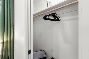 Un armario blanco con una barra de madera sosteniendo ganchos negros, un pulverizador blanco y un caja de almacenamiento gris.