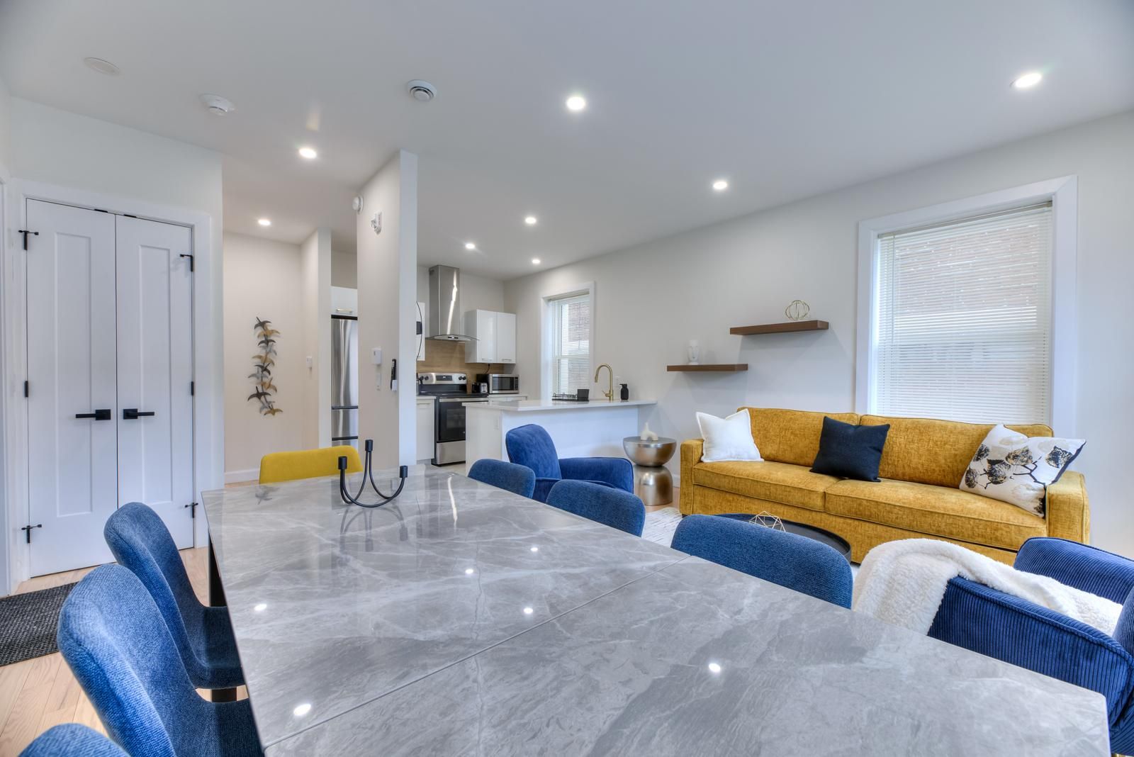 A modern living room with a marble table, blue chairs, a yellow sofa, and a kitchen in the background.