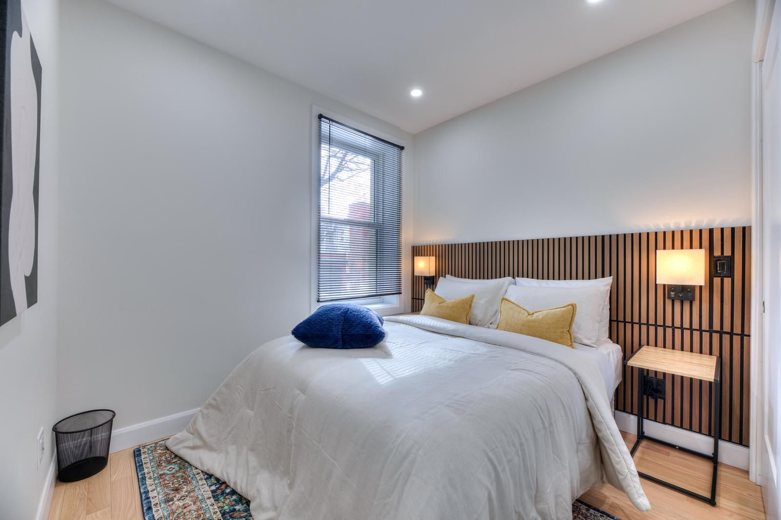 Une chambre à coucher moderne avec un lit, des lampes de chevet, une fenêtre avec des stores, une petite poubelle et un tapis à motifs.
