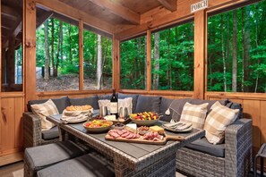 A cozy cabin interior with a dining table set for a meal, surrounded by wicker furniture and large windows offering a forest view. - Sleeps 8, Private Mini Golf, Hot Tub, Arcade, Log Cabin, 9 Mi to Dollywood (Sevierville)