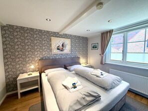 A hotel room with a double bed, two bedside tables, a nightstand, a window with curtains, and a wall with a patterned wallpaper. - North Sea Room 03 (Nieblum)