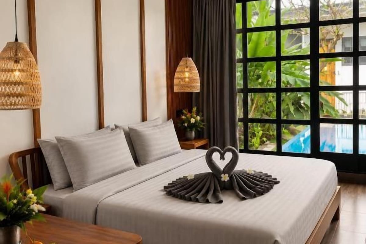 A modern bedroom with a large bed, a dining table with fruit, and a view of greenery through the window.