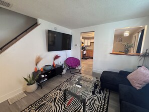 A modern living room with a flat-screen TV, a black entertainment unit, a patterned rug, a dark sofa, and a kitchen in the background. - Charming 4-bedroom duplex (Manchester)