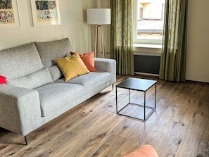 A living room with a grey sofa, a small black table, wooden flooring, a floor lamp, and a window with curtains. - Maloja Alpine Lodge by Interhome (Maloja)