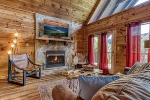 L’intérieur chaleureux d’un chalet avec une cheminée en pierre, des murs en bois, un téléviseur fixé au-dessus de la cheminée et un coin salon confortable avec un canapé et des fauteuils.
