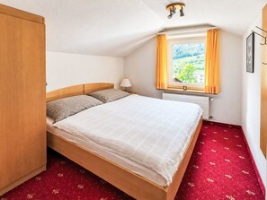 Een eenpersoonskamer met een houten hoofdeinde, een raam met gordijnen, een radiator en een rood tapijt.