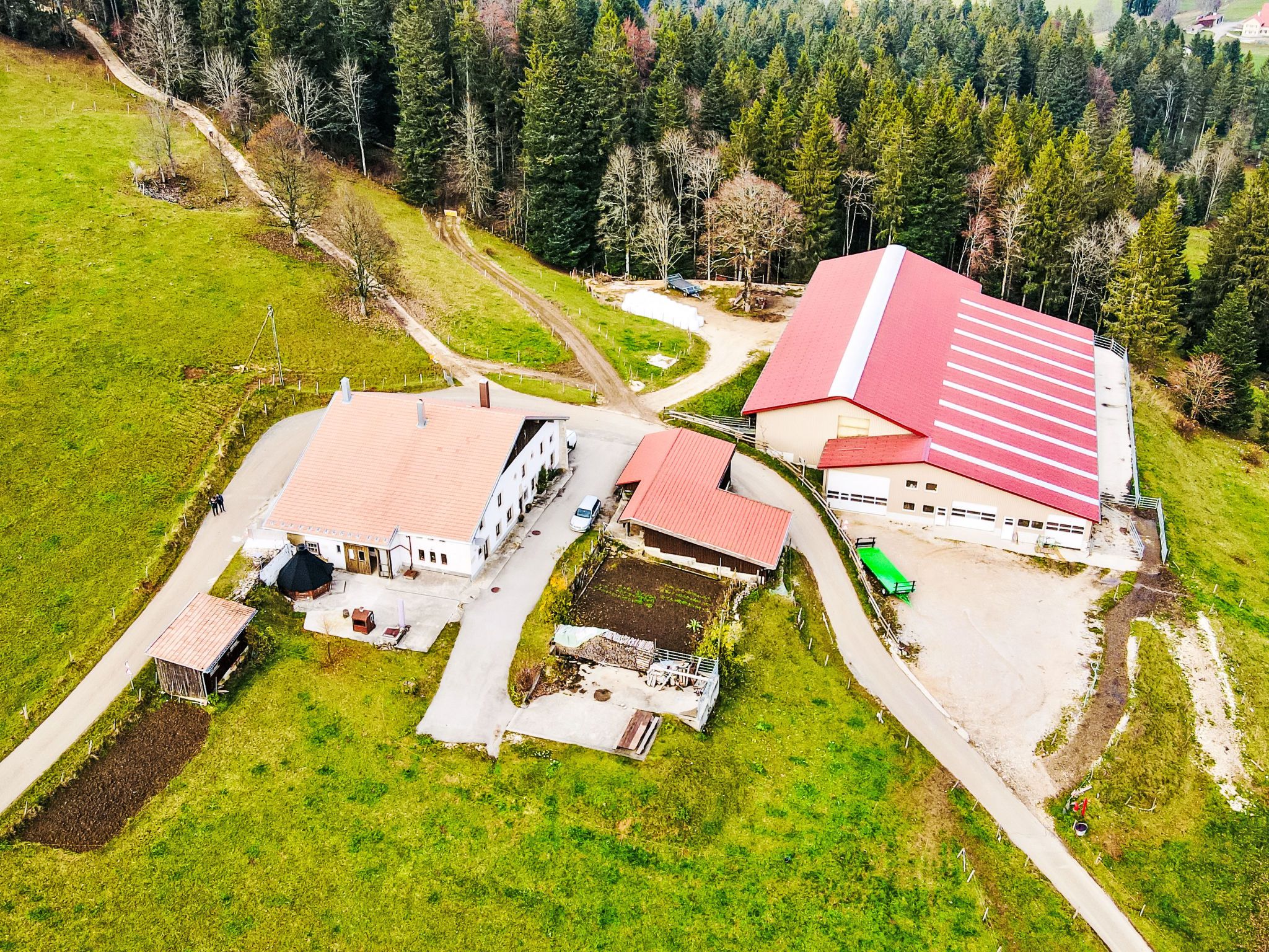 Vista aérea de una granja con varias construcciones, incluyendo un granero con techo rojo, rodeada de campos verdes y árboles.
