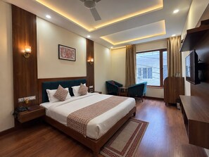 A hotel room with a large bed, a desk, a chair, a television, and a window with a view of buildings. - The Abhyudyam Ganga (Haridwar)