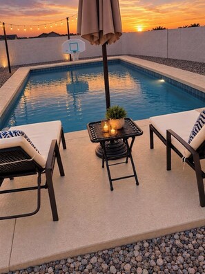 A swimming pool with a basketball hoop, two lounge chairs, a table with a plant and candle, and a sunset in the background. - Private Arcadia-Phoenix Escape with Resort-Style Backyard (Phoenix)
