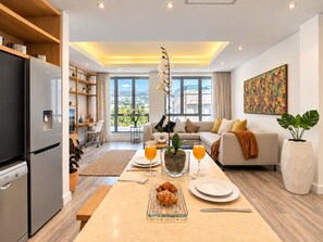 Cocina moderna con refrigerador, mesa de comedor con alimentos para el desayuno y sofá en la sala de estar con vista a la ciudad.