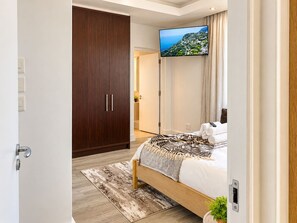 Inviting Bedroom Retreat: A beautifully furnished bedroom featuring a cosy wooden bed, elegant patterned throws and a spacious dark wood wardrobe. Soft lighting, a wall-mounted TV and contemporary finishes create a relaxing atmosphere. - New prospect Mandela Rhodes apartment (Cape Town)