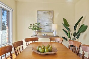 Ein Essbereich mit Holztisch, Stühlen, einer Schüssel mit Birnen, einer Vase mit Blumen, einer Lampe und einem großen abstrakten Gemälde an der Wand.