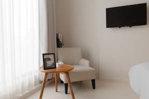 A minimalist room with a white chair, a small wooden table with a picture frame, and a flat-screen TV mounted on the wall. - Digital Nomad Studio Room Kuta (Badung)