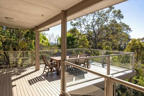 Une terrasse en bois avec une rambarde en verre, des meubles d’extérieur et une vue sur les arbres et le ciel.