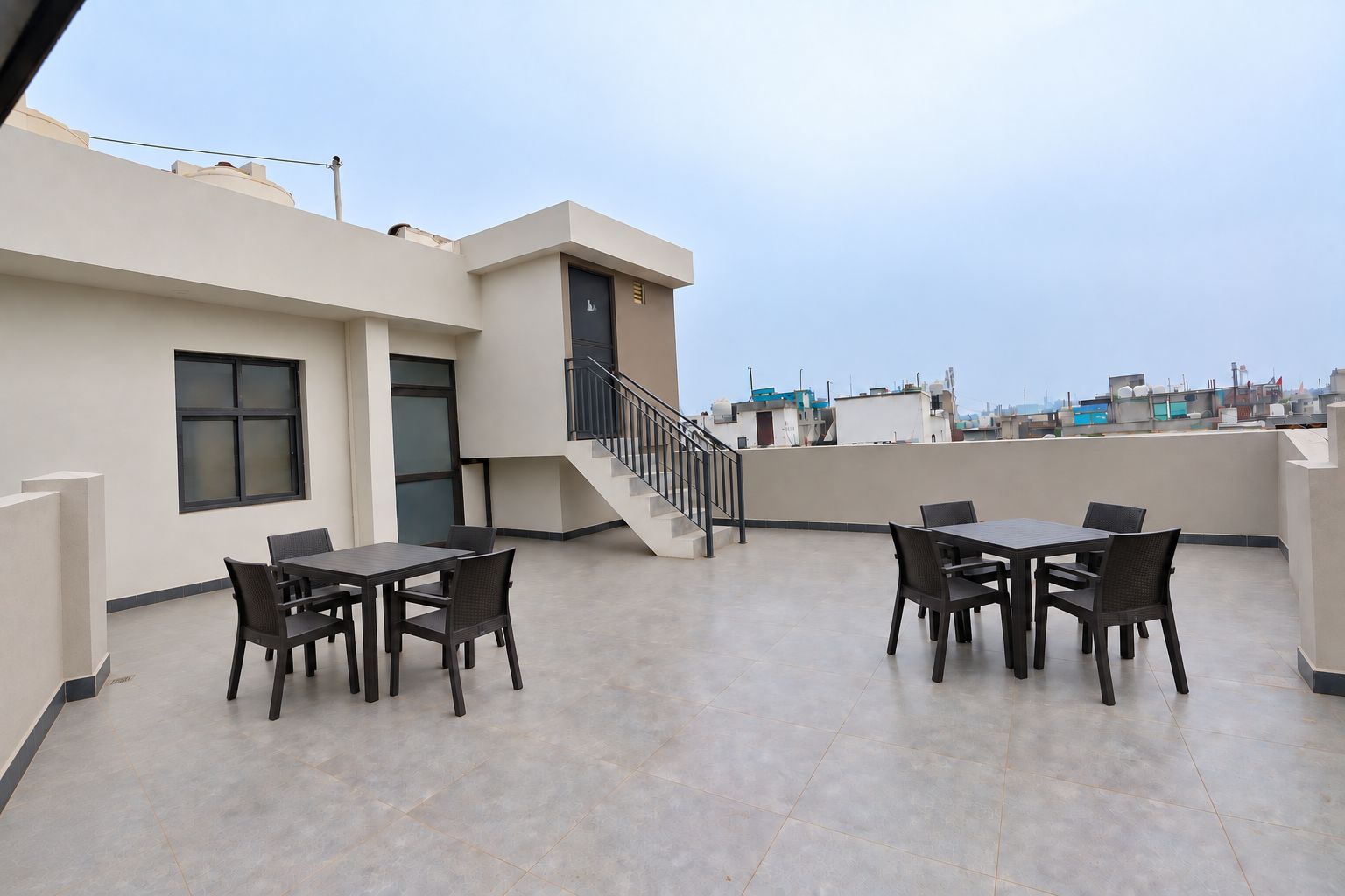 A rooftop terrace with two tables and chairs, a staircase, and a view of the cityscape.