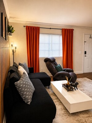 Ein Wohnzimmer mit einer schwarzen Sofagarnitur, einem weißen Couchtisch und orangenen Vorhängen.