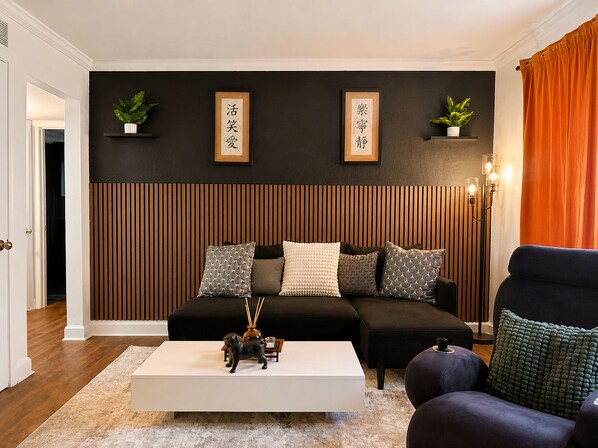 A modern living room with a black sofa, white coffee table, and a dark armchair. There are two framed calligraphy pieces on the wall, a floor lamp, and a potted plant.