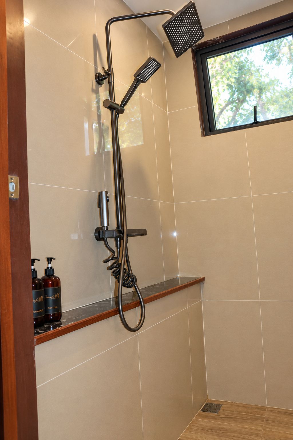 Une douche équipée d’un pommeau de douche à main et d’un pommeau de douche fixe, une étagère en bois pour les articles de toilette et une fenêtre donnant sur les arbres.
