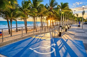 Een rolstoeltoegankelijk pad langs een strandboulevard met palmbomen en een ondergaande zon.