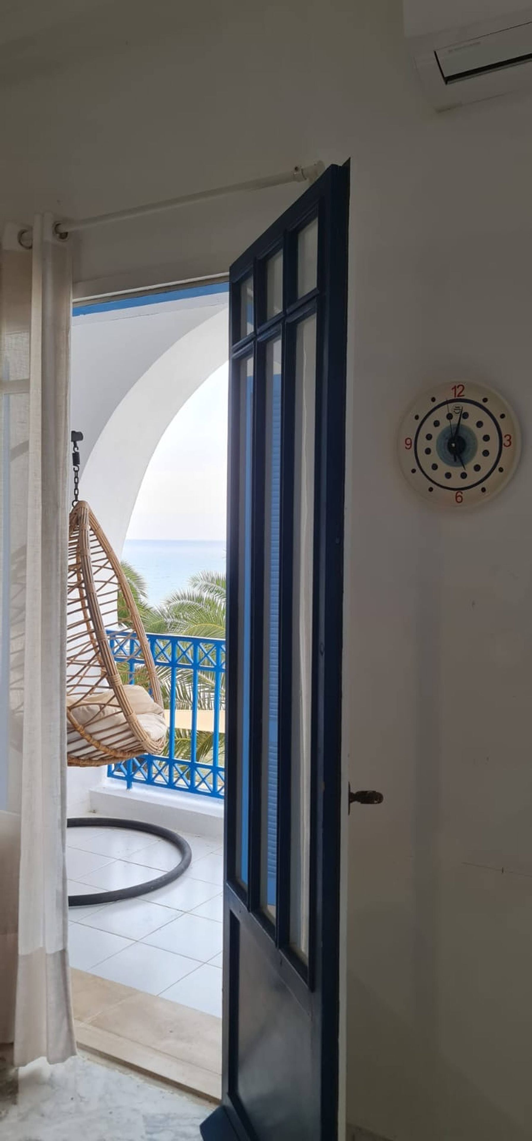 Une porte ouverte donnant sur un balcon où se trouve un fauteuil suspendu, une rambarde bleue et une horloge murale.