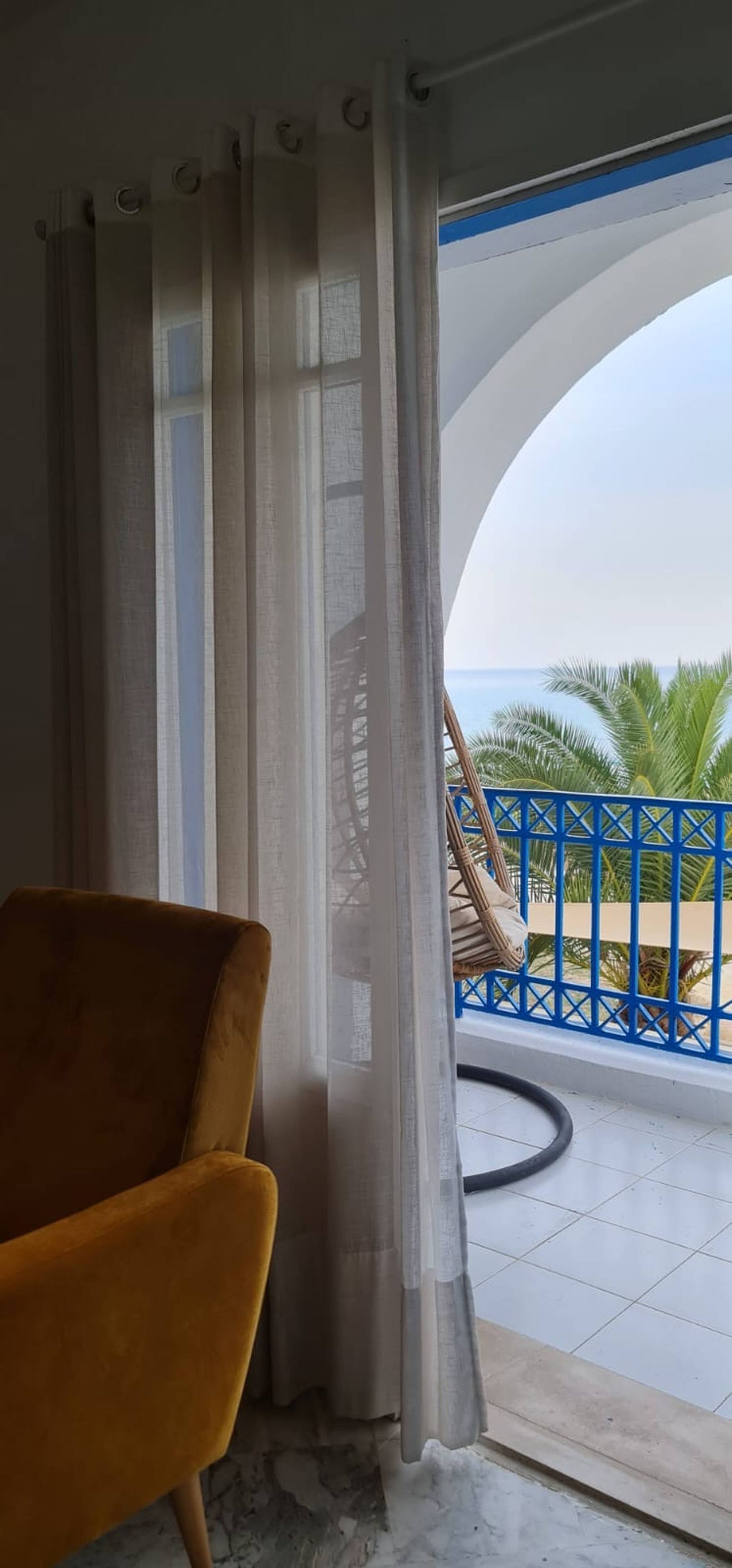 A room with a view of the sea, a chair, and a balcony with a blue railing.