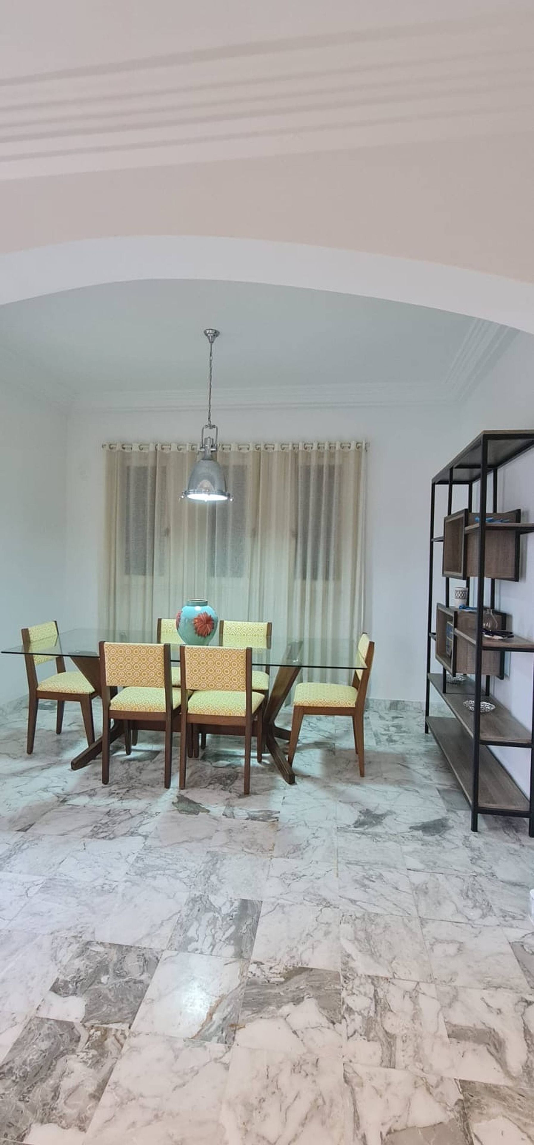 Un coin repas avec une table et des chaises en verre, un luminaire suspendu, un sol en marbre et une bibliothèque.