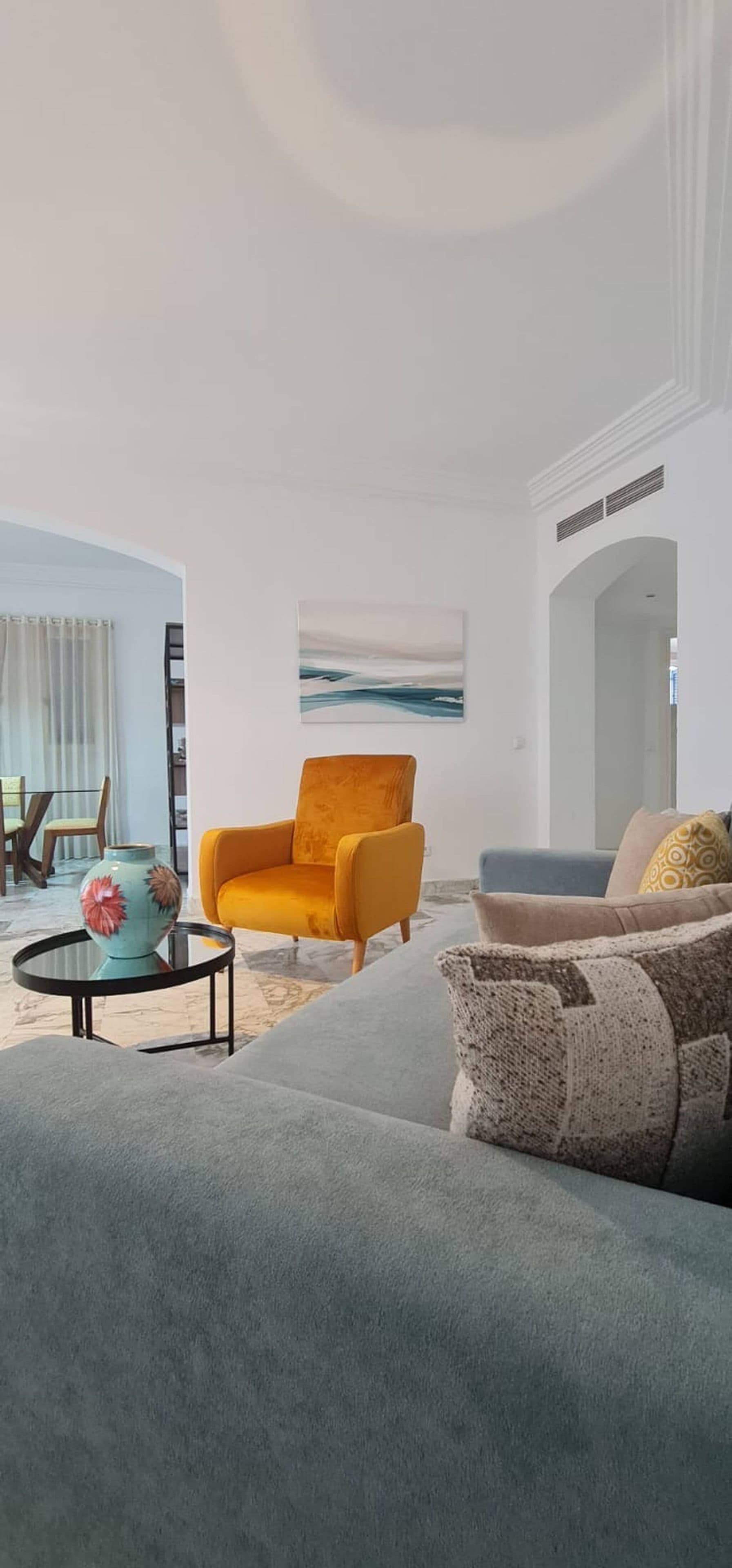 Un salon moderne avec un canapé gris, un fauteuil jaune et un canapé bleu. Un tableau est accroché au mur et un vase est posé sur une table.