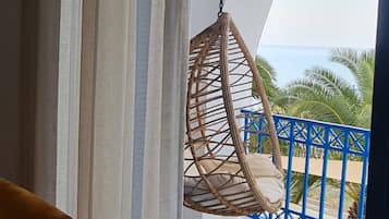 Une pièce avec un fauteuil suspendu, un fauteuil jaune et une vue sur la mer, accessible par un balcon doté d’une rambarde bleue.