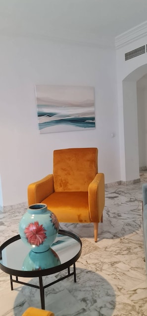Quarto com piso de mármore, uma poltrona laranja, uma mesa redonda com um vaso e um quadro de uma paisagem na parede.