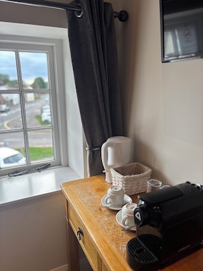 A room with a window, a wooden table, a white coffee maker, a basket, and a black bag.