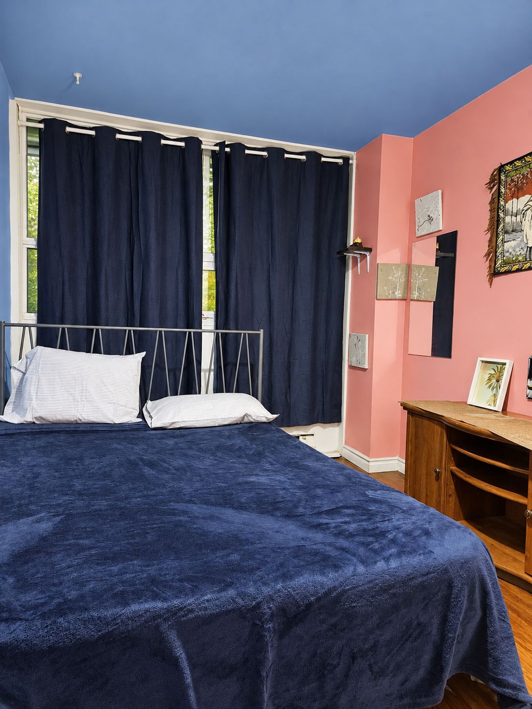 A bedroom with a bed, a window with curtains, a wall with artwork, and a wooden cabinet.