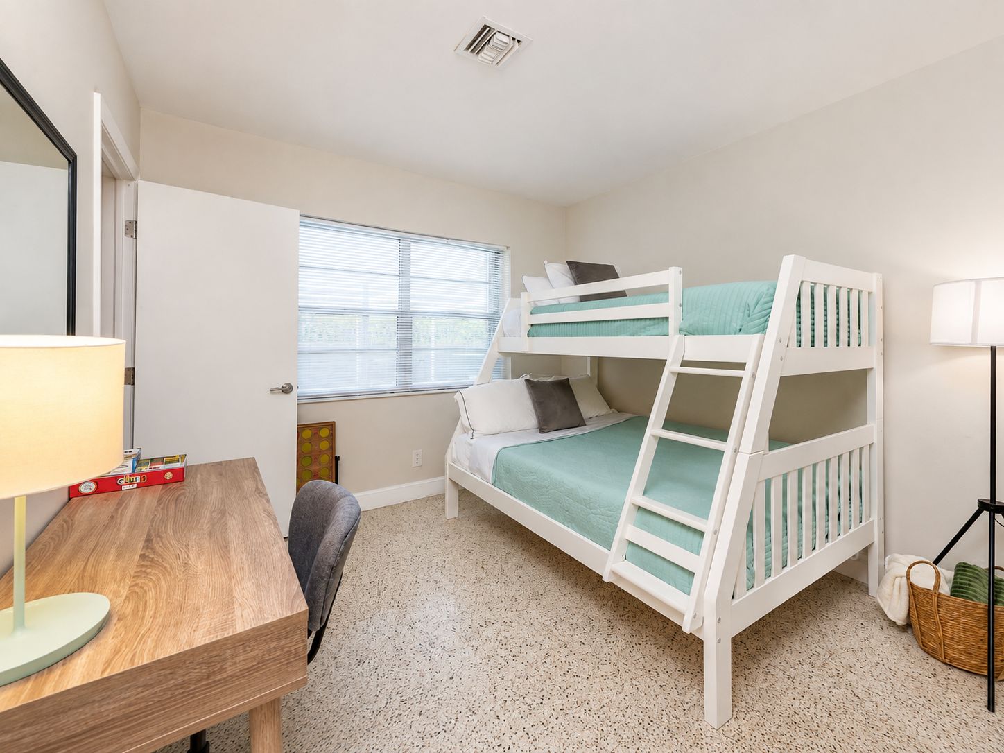 A bunk bed with a desk and chair, a lamp, and a window with blinds.