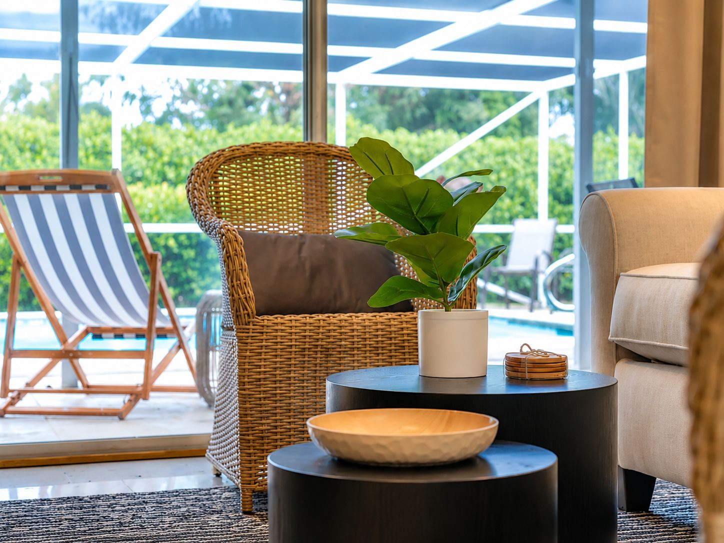 A wicker chair with a cushion, a potted plant, a bowl, and a side table in a room with a view of greenery.