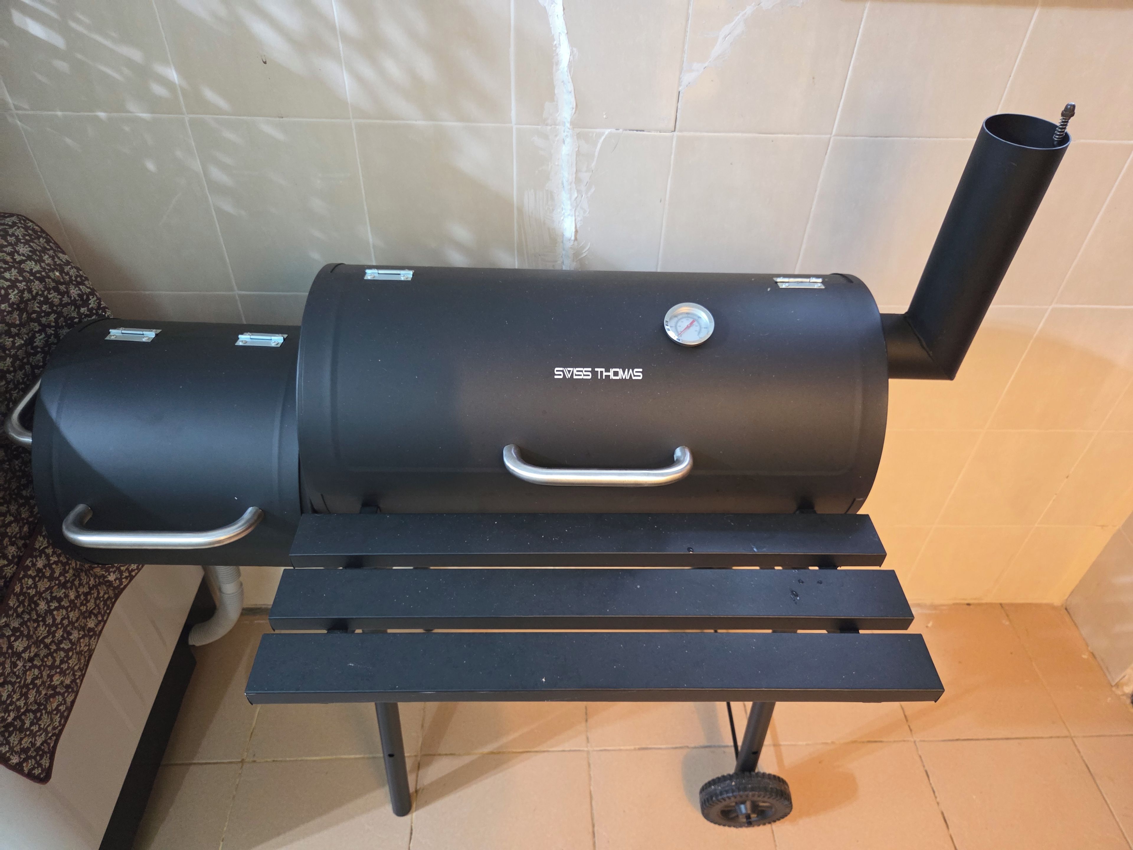 A black barbecue grill with a handle and a dial, placed on a tiled floor with a bench in front.