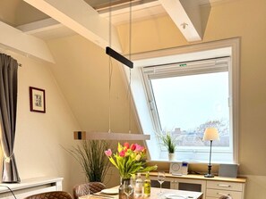 A dining area with a table set for two, a window with a view, and a ceiling with exposed beams. - "deichhof", No. 26 (Utersum)