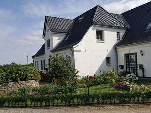 A two-story white building with a dark roof, surrounded by a green fence and a variety of flowering plants. - "deichhof", No. 26 (Utersum)