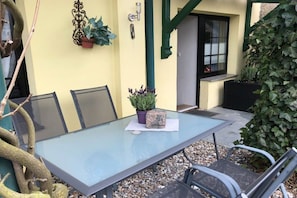 Et bord med glasplade og fire stole, en potteplante og et velkomstskilt på en terrasse med gul væg og grønne detaljer.