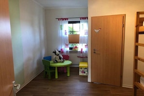 Et lille rum med en trædør, et vindue med gardiner, en grøn stol, et lille bord og en bamse.