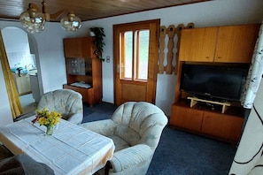 Ett rum med en tv, ett matbord med blommor, två fåtöljer, ett träskåp och en dörr med ett fönster.
