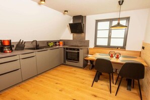 A compact kitchen with a built-in oven, a coffee maker, and a dining area with a table and chairs. - Zugspitze Apartment Panoramic View (Altenberg)