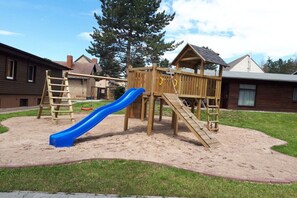 A playground with a slide and climbing structures, surrounded by sand and grass, with houses in the background. - Langenthal Forestry House - House II (Braunsroda)