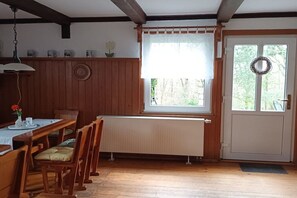 Een eetgedeelte met een houten tafel, stoelen, een radiator en een raam met uitzicht op bomen.
