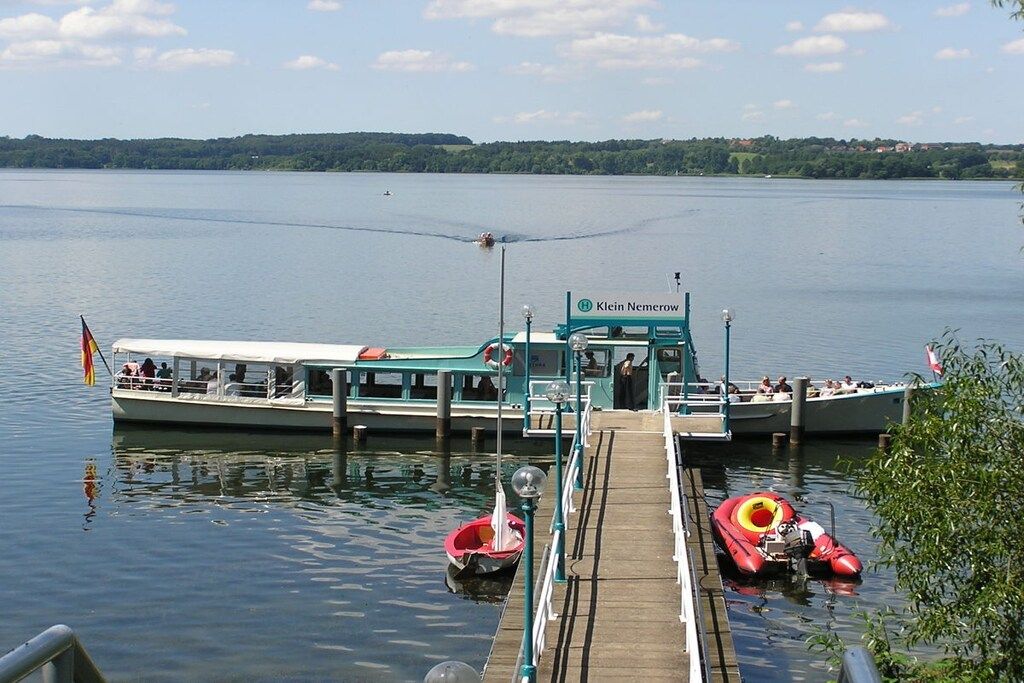 Ein Boot liegt an einem Steg mit Blick auf einen stillen See und eine bewaldete Uferlinie.
