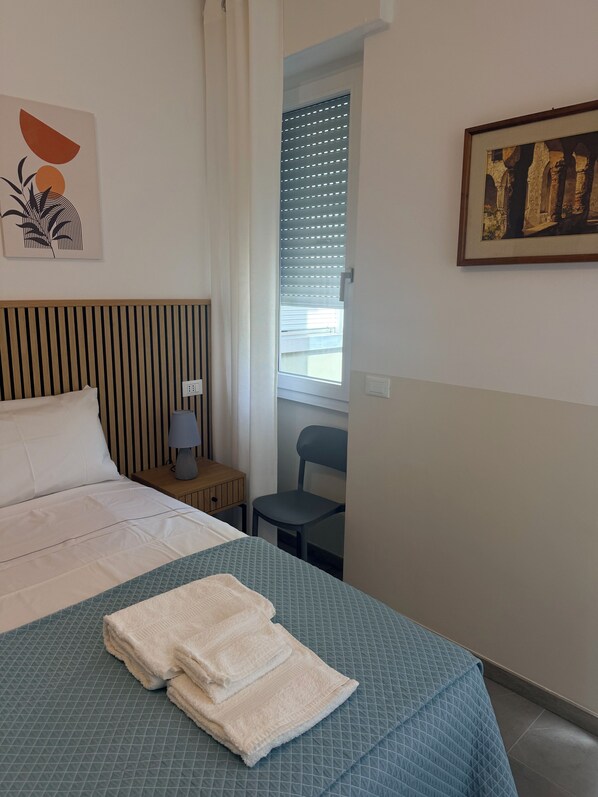 Una habitación de hotel con cama, silla, mesita de noche, lámpara y ventana con persianas.