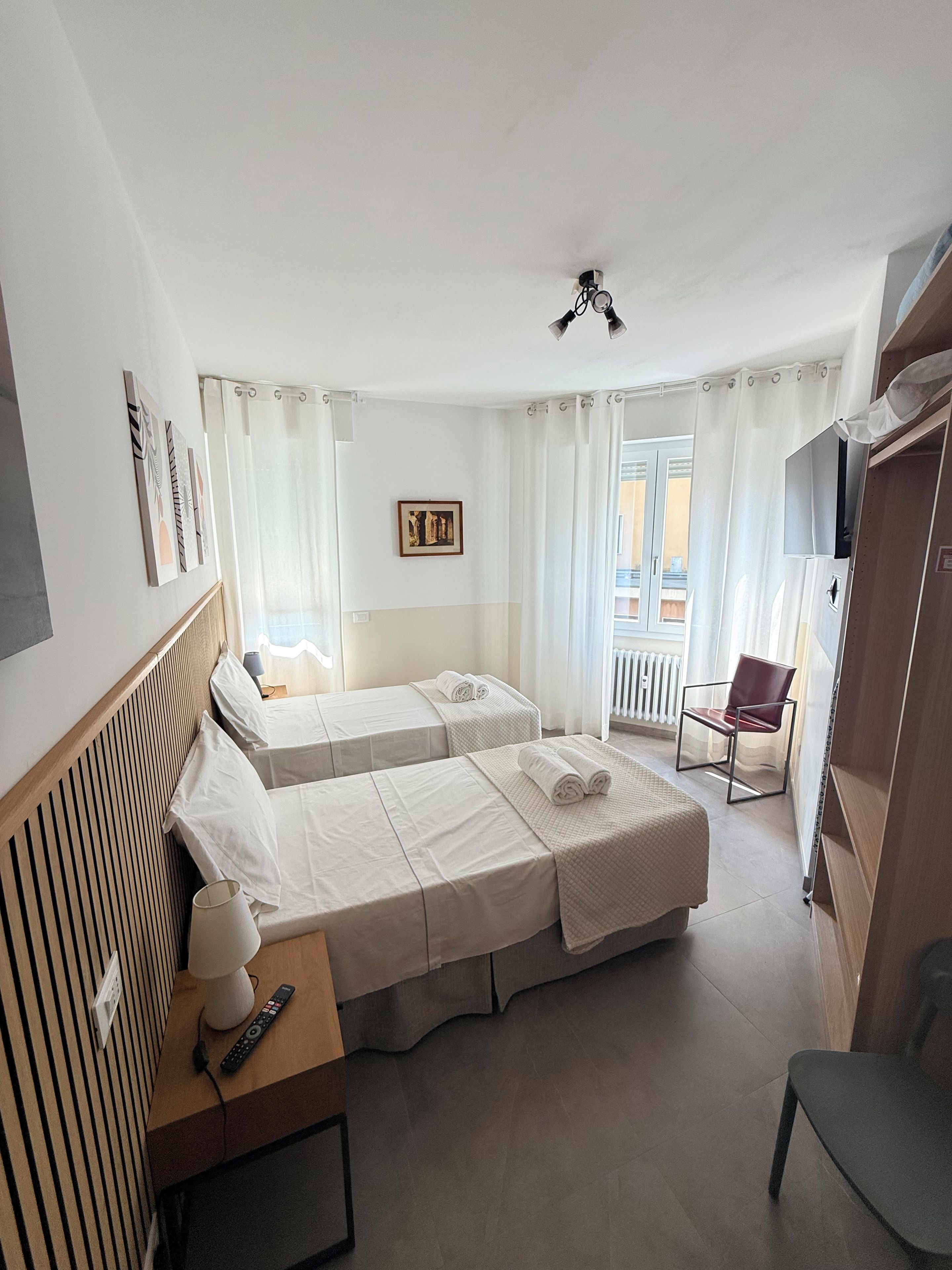 Una camera d'albergo con due letti, una sedia, una scrivania con lampada, una televisione e una finestra con tende.