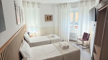 Una camera d'albergo con due letti, una sedia, una scrivania con lampada, una televisione e una finestra con tende.