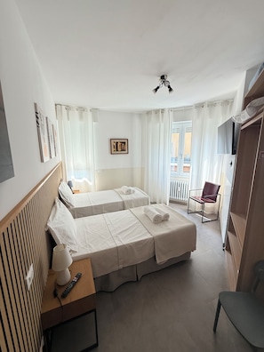 Habitación de hotel con dos camas, una silla, un escritorio con lámpara, televisión y ventana con cortinas.