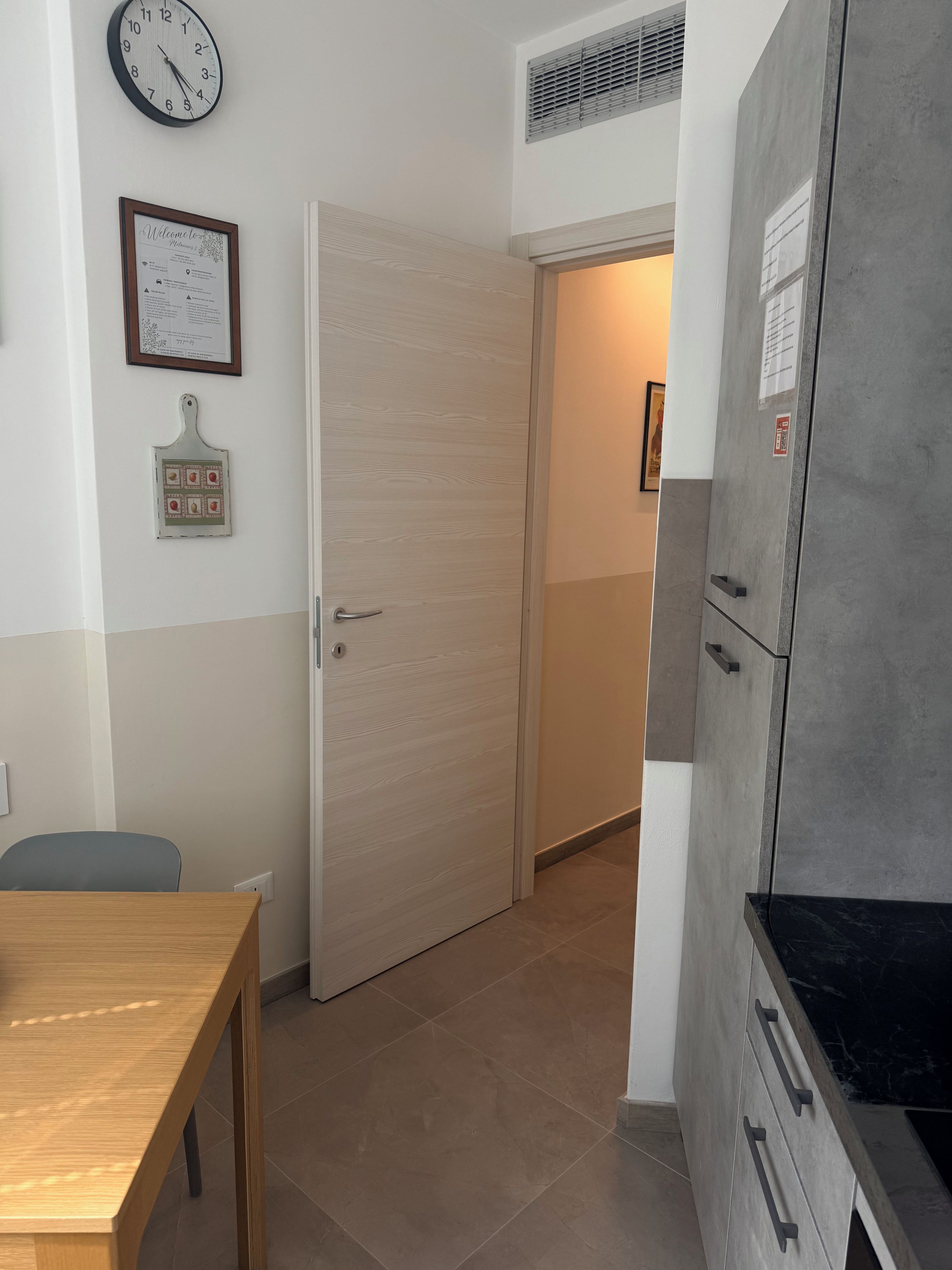 Una stanza con un tavolo in legno, una porta, una sveglia a parete e un frigorifero.