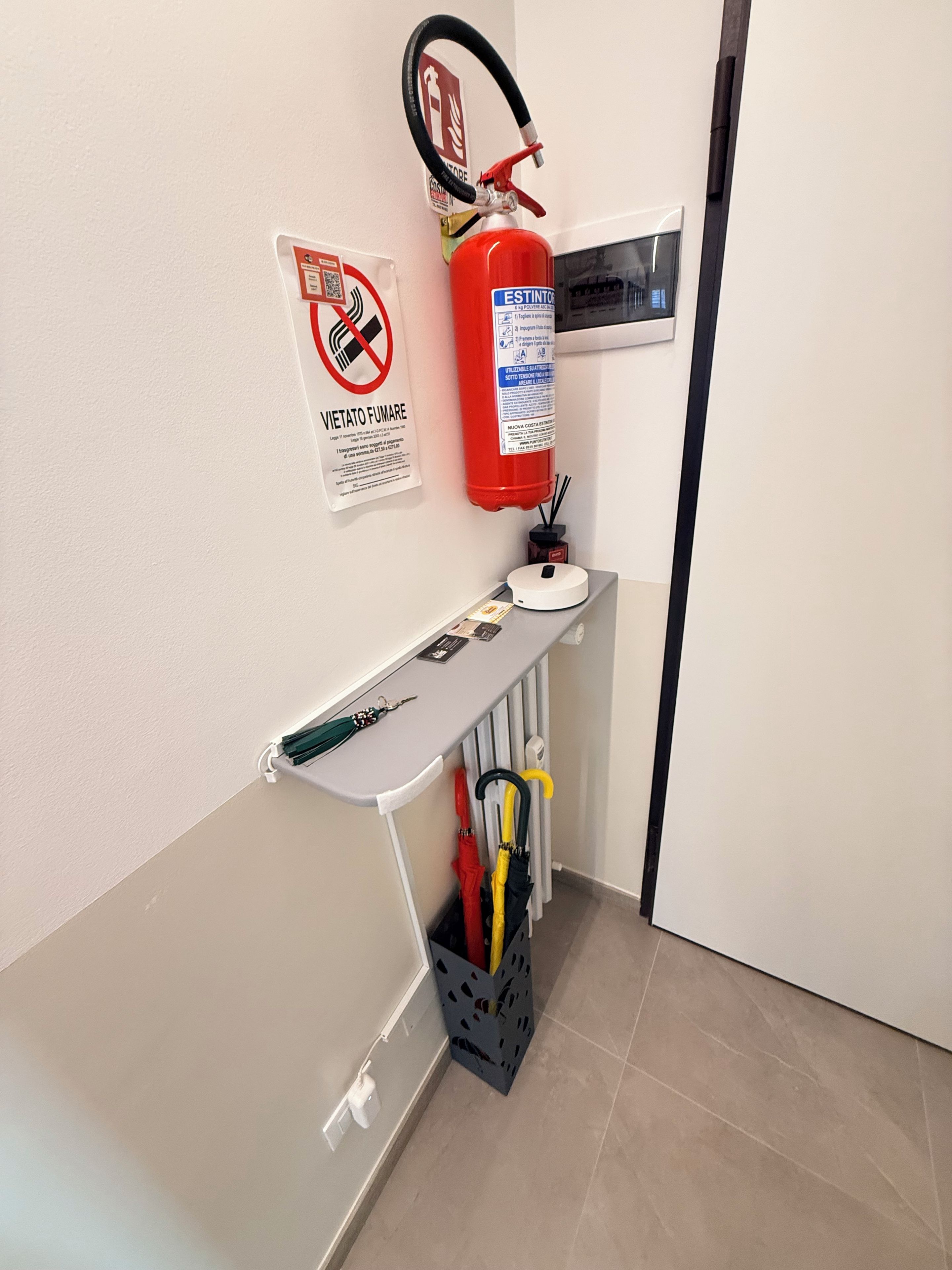 Una stanza con un estintore fissato al muro, un tavolino con un vassoio e un portaombrelli.