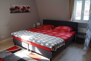 A bedroom with a bed, a nightstand, a radiator, and a window with curtains. - Holiday apartment with 1 bedroom (Königstein)